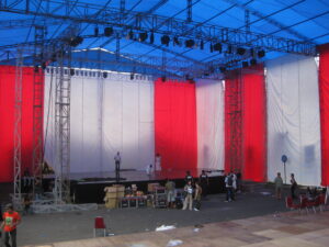 load in for a circus show. blue tent ceiling and red and white walls surround a truss. crew members work on and around the stage.