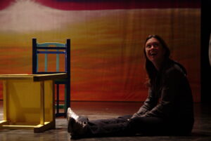 Mia Caress sits with legs extended on the ground. Two chairs, part of a circus set, are nearby. The background is a fade of orange and white.
