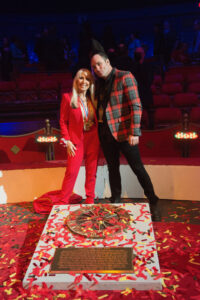 At the 2022 Circus Ring of Fame Awards, American acrobat "Galaxy Girl" Tina Winn, a blonde woman in a red pantsuit, stands over her Circus Ring of Fame plaque to commemorate her career achievements