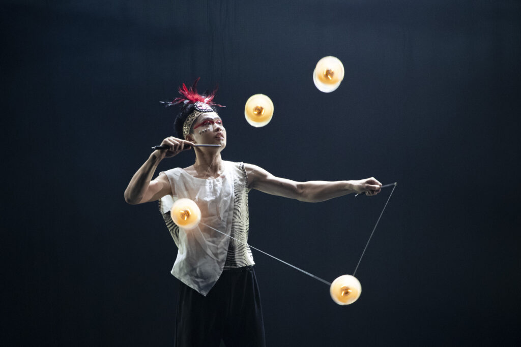 Taiwanese performer Peng Chan, wearing a red feathered headdress, does tricks with 4 diabolos on stage