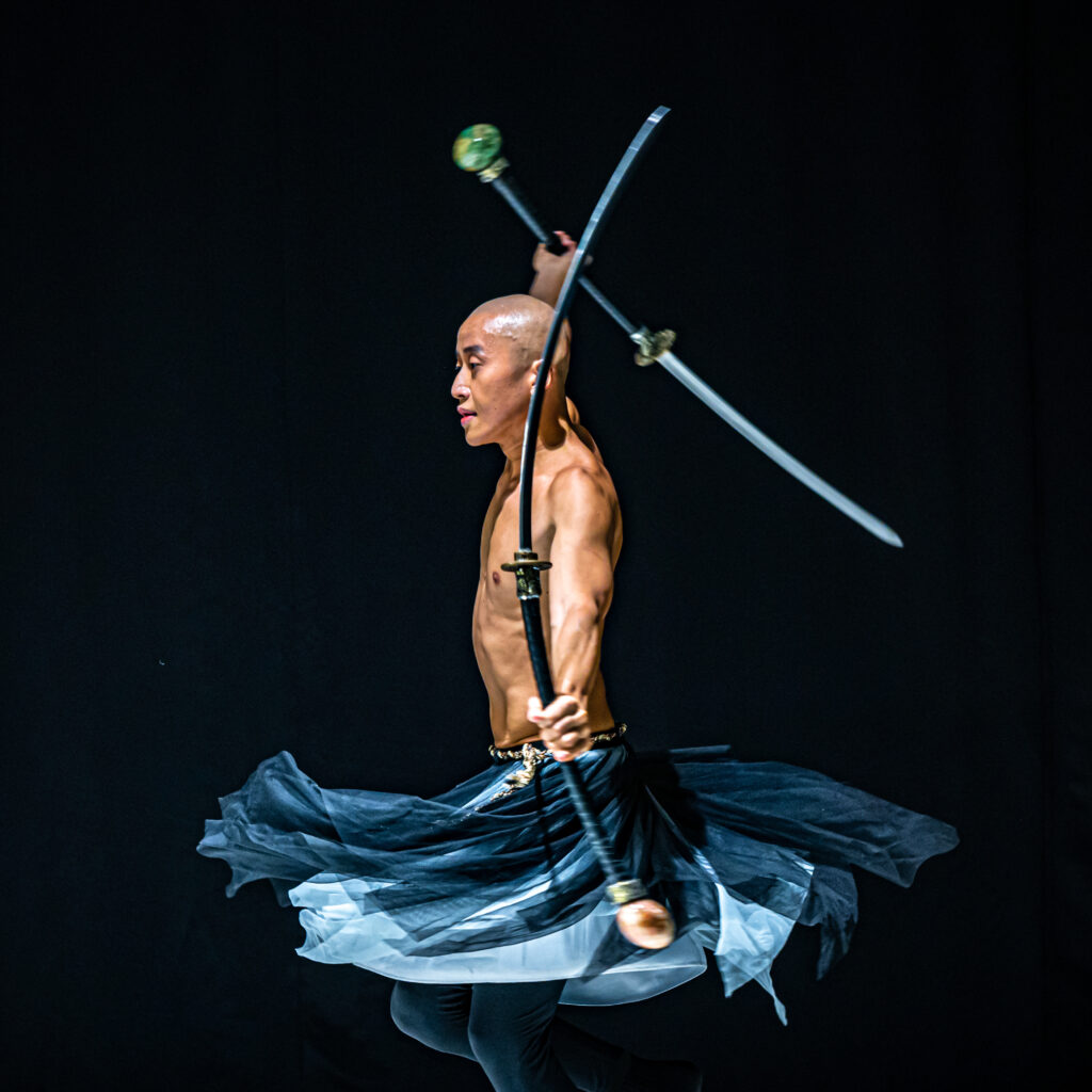 Taiwanese sword dancer and "contact sword" handler Titos Tsai performs his own act, "Flaming Sword." Tsai is a bald and shirtless man who holds two decorative swords