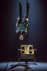 Circus performer Baptiste Clerc hangs upside down above a prop chair on a dark stage backdrop