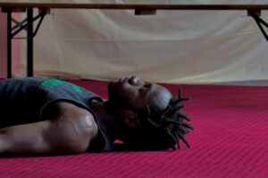Acrobat Antino Pansa, a young Black man with short dreadlocks in a sleeveless shirt, rests on his back after a training studio session
