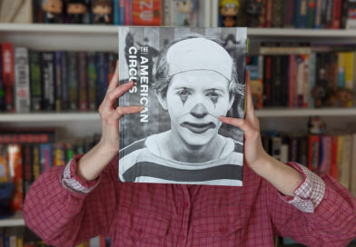 In front of her bookshelf, Morgan Anderson holds up a copy of the book The American Circus by Susan Weber et. al. over her own face