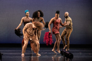 Six circus performers dressed in shades of brown dance and whirl their hair while holding colorful wigs. 