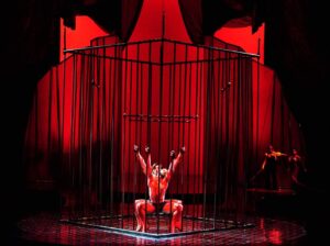 Part of Cirque du Soleil's adult circus show Zumanity, Arnaud Boursain, wearing red stilettos and a costume, does an acrobatic dance-squat within a large cage 