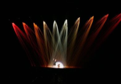 A single performer in center stage, illuminated under ten spotlights; the outer eight are red lights.