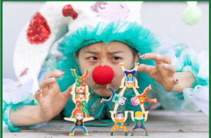 Wearing a red clown nose, a young Asian girl with a focused expression builds pyramids out of toy circus performers