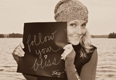 Circus consultant and CircusTalk CEO Stacy Clark, a smiling blonde Canadian woman wearing a beanie, holds a sign with "follow your bliss" written in cursive while standing on the shores of a local river