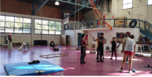 Inside of a municipal sports complex in Chile, a group of circus performers from the Circo Hermanos Castro stands and converses between trainings. Mats and scattered acrobatic equipment fill the space around them