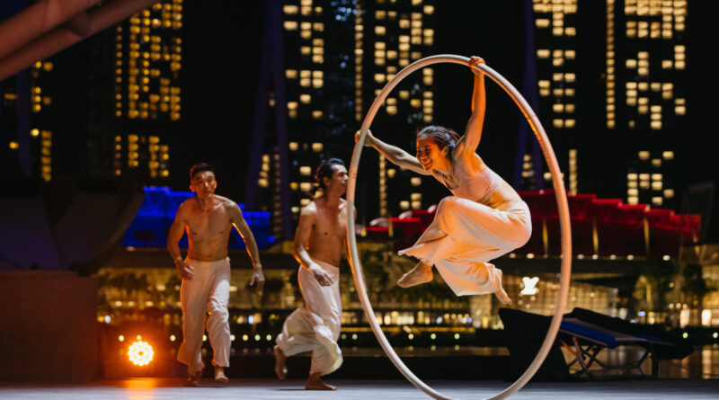 Three Bonfire Circus performers, two young Asian men and a female aerialist in a Cyr wheel, on an outdoor stage at the circus festival Flipside 2023. Some of Singapore's iconic skyscrapers lit up at night are visible in the background.
