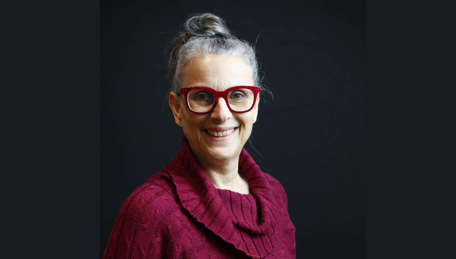 Nadia Drouin, Executive Director of En Piste, is a middle-aged woman with gray hair in a bun, a smile, and smart attire. She wears a red cowl-necked sweater that matches her red-framed glasses.