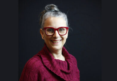 Nadia Drouin, Executive Director of En Piste, is a middle-aged woman with gray hair in a bun, a smile, and smart attire. She wears a red cowl-necked sweater that matches her red-framed glasses.
