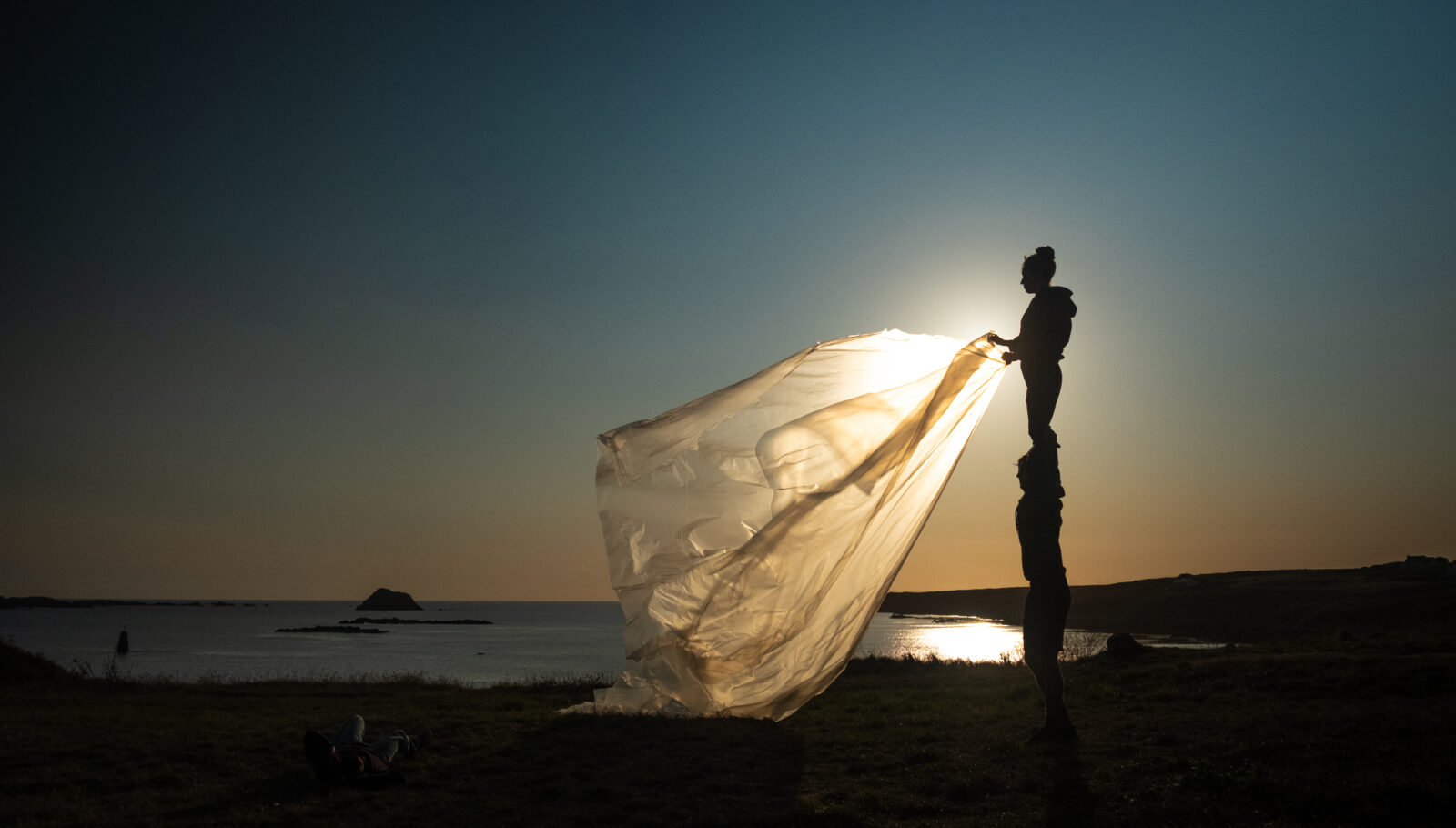 Circus Barcode artist Alexandra Royer in performance on a beach, part of her work with "Elementerre". She stands in profile silhouette with one side to the sunset, unfurling a billowing plastic sheet with both hands